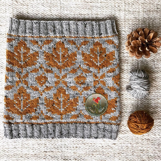 Knitted cowls with autumn leaf pattern on a textured surface with pine cones and yarn.
