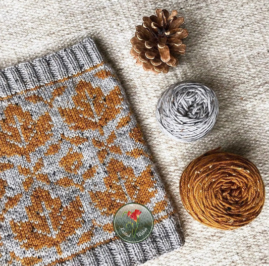Knitted cowl with orange and gray maple leaf colorwork, yarn ball, pine cone on textured surface
