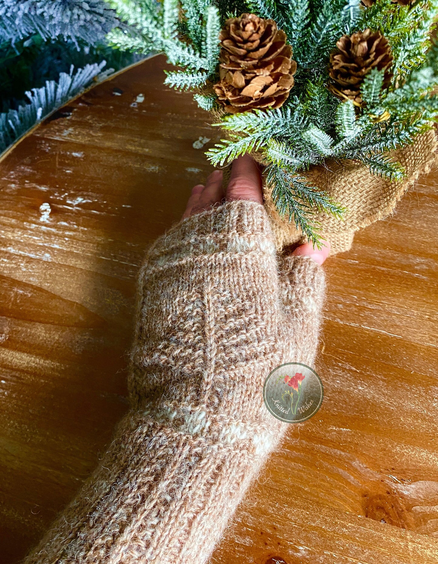 Hand wearing a textured knitted mitt holding a small Christmas tree on a wooden surface.