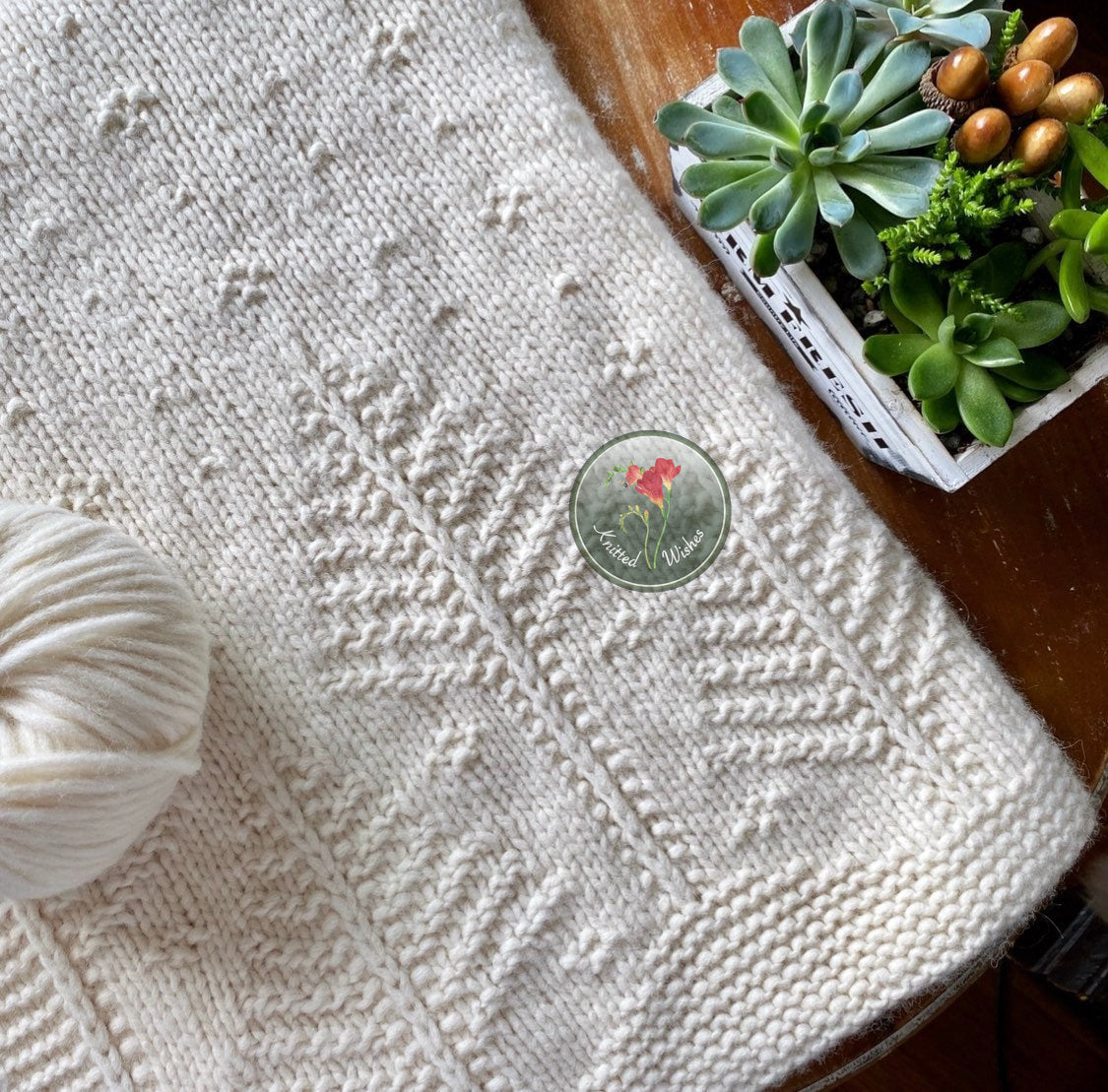 Knitted blanket with a tree pattern, yarn ball, and potted succulents on a wooden surface