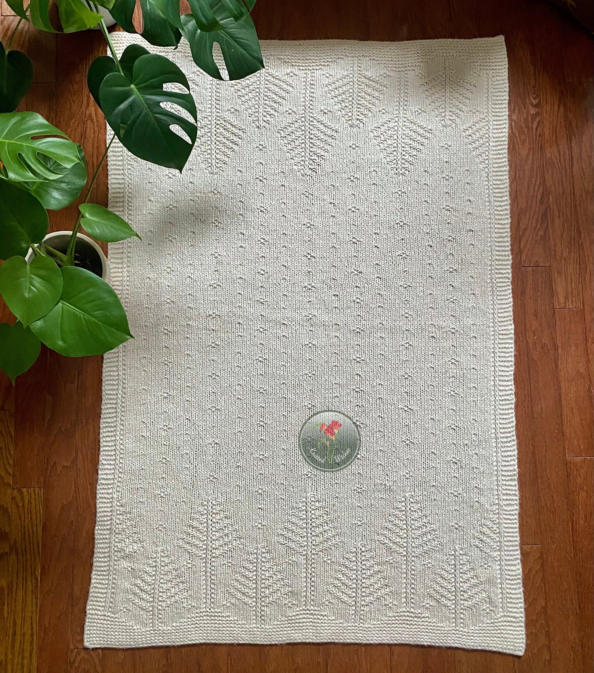 White rectangular textured knitted tree blanket on a wooden floor next to a plant.