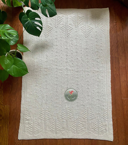 White rectangular textured knitted tree blanket on a wooden floor next to a plant.