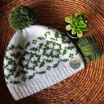 Knitted colorwork white hat with green floral pattern and pom-pom on a woven surface with a small plant.